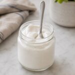Glass jar of high protein marshmallow fluff with a spoon standing upright
